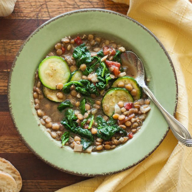 Green bowl of soup with vegetables on a wooden table with bread and a yellow napkin.