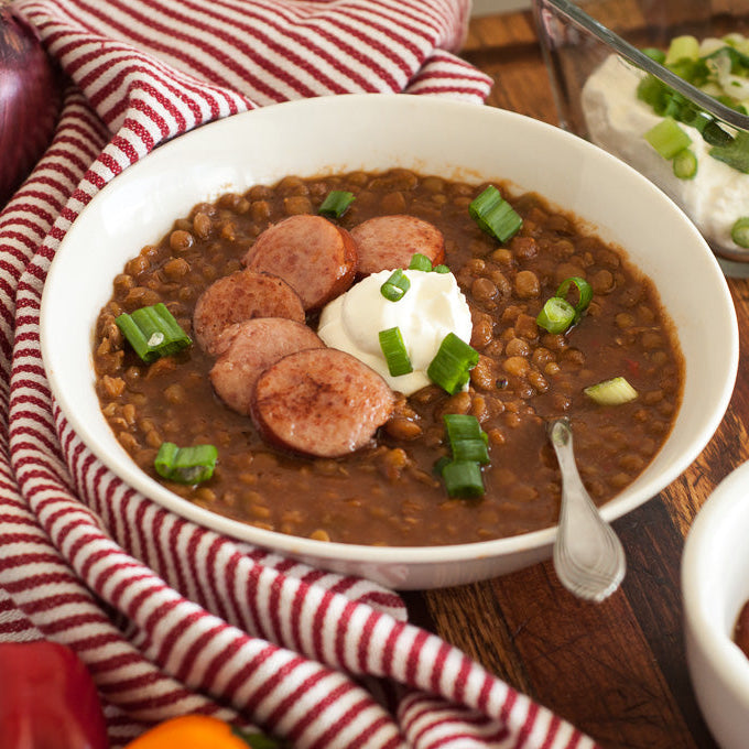 Bowl of chili with sausages and garnishes on a wooden table with a striped cloth.