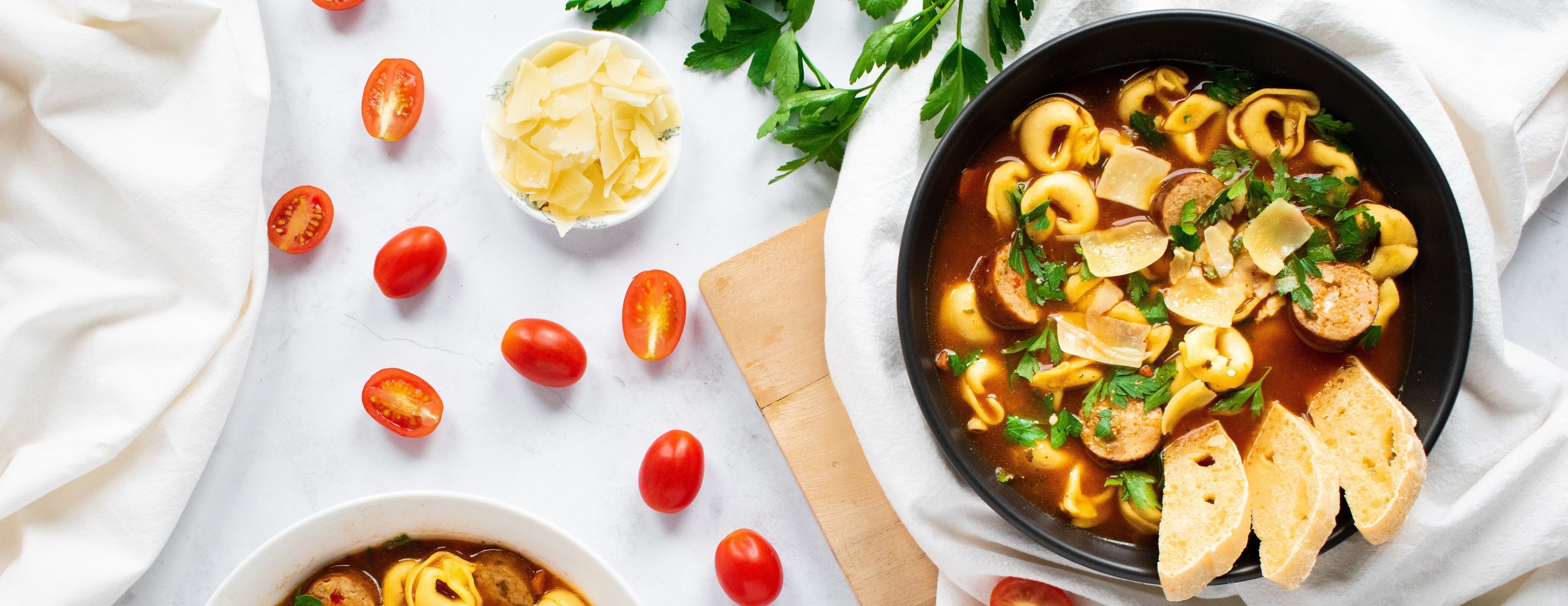 Two bowls of tortellini soup with cherry tomatoes and herbs on a white surface.